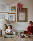 Two young children sit at a wooden table making cookies, with flour, baking tools, and the Mushie Bluey x Mushie Silicone Training Cup + Straw nearby. Framed art and a wall lamp in the background create a cozy atmosphere.