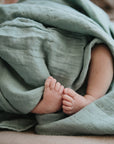 A close-up of a baby’s bare feet peeking out from under a soft, green mushie Organic Cotton Muslin Swaddle Blanket.