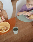Two young children eat pasta and veggies from divided plates, each using a fork and sipping from mushie Dinnerware Cups. One holds a cucumber slice. The scene is bright and casual.