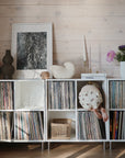 A toddler climbs into a white shelving unit filled with vinyl records, while a mushie Snack Cup sits nearby. The shelf holds vases, framed art, a candle, and flowers. Light wooden panel walls create a cozy, modern vibe in the room.