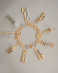 A flat lay of mushie’s beige Flower Training Toothbrush, pacifiers, bottle, spoons, forks, clips, and nipple soothers arranged in a circular sunburst pattern on a white background.