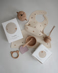 Flat lay of beige and light brown baby feeding and teething items, plus two boxed mushie Organic Cotton Muslin Swaddle Blankets.