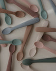 Mushie Silicone Feeding Spoons, made from food-grade silicone, are displayed on a white surface in soft pastel shades of pink, blue, green, and beige. Each spoon features the “mushie” logo embossed on the handle.