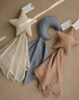 Three mushie Moon Lovey Blankets—organic cotton comforters—rest on beige: a cream star, a blue crescent moon and cloth, and a dusty pink star. Each is the perfect baby security blanket.