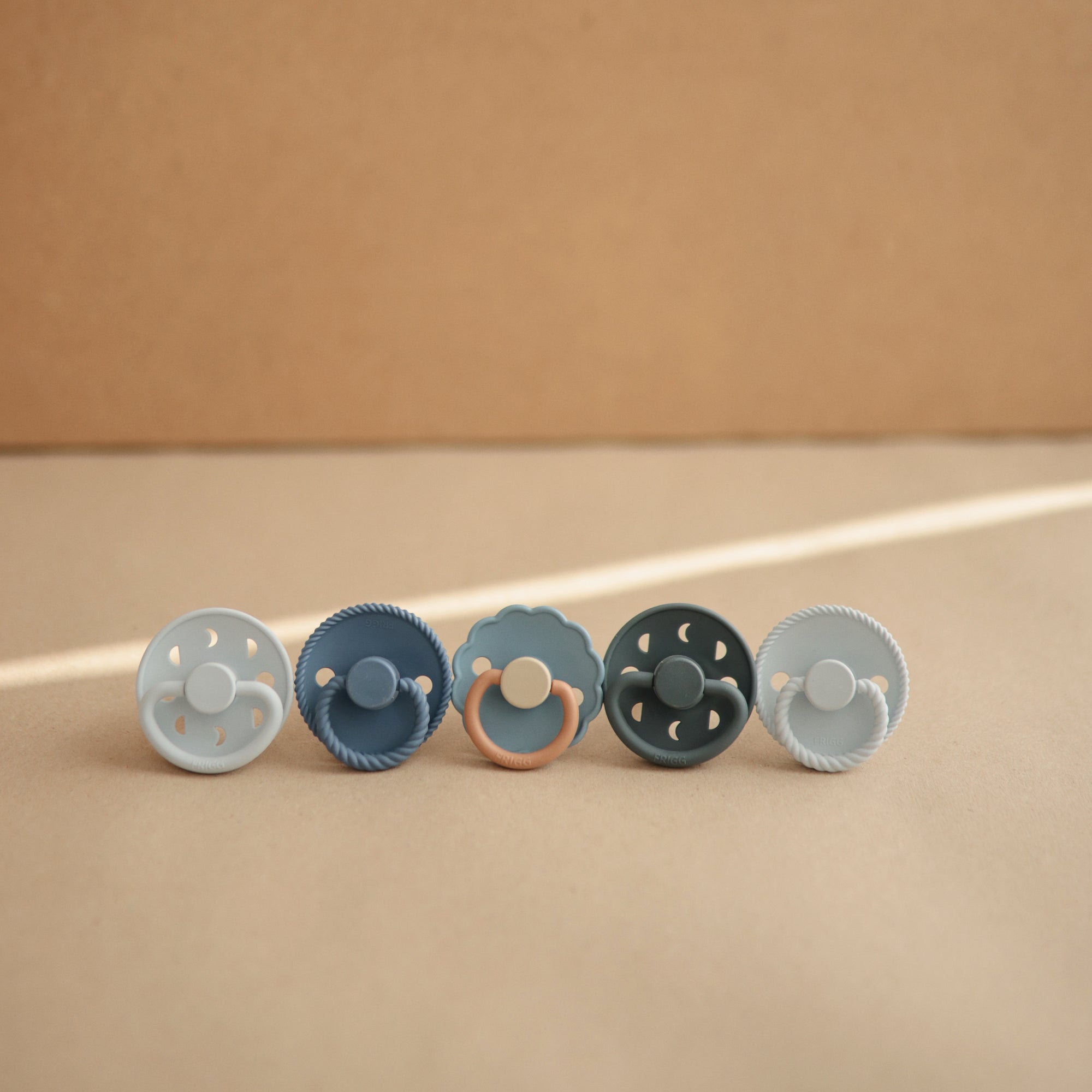 Lifestyle image of a blue-themed line of Frigg pacifiers, featuring the Powder Blue and Slate Moon Pacifiers.