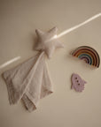 A mushie Star Lovey Blanket, a pastel rainbow stacker, and a lavender rocket toy are arranged on a beige surface with sunlight streaming diagonally.