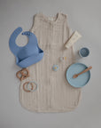 A beige baby sleep sack is displayed with a blue silicone bib, baby lotion, pacifier, wooden teether, beaded teething ring, blue bowl with spoon, and mushie Dinnerware Cups on a light grey background.