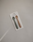 A set of two mushie Silicone Feeding Spoons, in dark green and light brown, shown on a white backing against a plain light gray background.