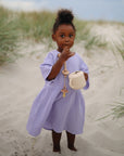 A young child in a light purple dress stands on sandy dunes, holding a mushie Snack Cup and a pacifier clip, looking at the camera with one hand near her mouth. Another child is partially visible on the left.