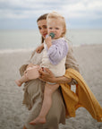 A woman stands on a sandy beach holding a child with a pacifier and pink watering can. A mushie Organic Cotton Muslin Swaddle Blanket is draped over her arm, set against the sea and a cloudy sky for a serene scene.