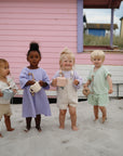 Four toddlers, barefoot on sand in front of a pink beach hut, each hold a mushie Snack Cup like a toy purse. They wear summery outfits and smile or look curiously at the camera, with pastel-colored huts completing the scene.