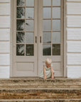 A young child in a light dress sits on stone steps outside a white brick building, holding the FRIGG Andersen Fairytale Silicone Pacifier 2-Pack and looking down as they touch the steps with both hands.