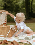 A young child with blonde hair sits on a blanket in a grassy park, reaching into an open picnic basket. A FRIGG Andersen Fairytale Silicone Pacifier from FRIGG rests nearby. Trees and greenery are visible in the background.