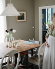 A pregnant woman stands in a softly lit dining room with a wooden table set with flowers, candles, and mushie Silicone Placemats made from food-grade silicone, surrounded by white chairs and muted green walls adorned with a painting.