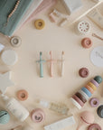 A flat lay of pastel mushie baby products, including the Flower Training Toothbrush, creams, stacking toys, teethers, skincare bottles, and soft cloths, all neatly arranged on a beige surface.