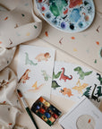 Art supplies and baby items with dinosaur patterns are displayed on a table, featuring dinosaur watercolor paintings, a palette, brush, bib, cloth, teething ring, and a mushie Silicone Placemat.