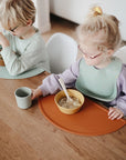 Two young children with light hair and glasses sit at a wooden table, eating from bowls on mushie Silicone Placemats. The child on the right reaches for a cup; both wear bibs and casual clothes, enjoying their meal together.