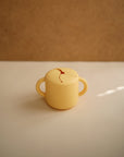 The mushie Snack Cup is a small, yellow, spill-proof silicone snack cup with two handles and a flexible slit lid—perfect for toddlers. It sits on a white surface against a brown background.