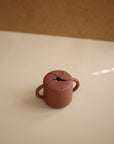 The mushie Snack Cup is a small, brown silicone snack holder with two handles and a spill-proof, petal-like lid—perfect for toddlers. It sits on a light surface against a beige background.