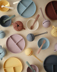 The mushie Snack Cup and matching silicone baby plates, bowls, and spoons in soft pastels are arranged on a light surface. Some plates feature animal faces and divided compartments, offering a cute and practical mealtime set for toddlers.