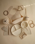A flat lay of beige and white mushie baby feeding accessories, including a spill-proof Snack Cup, bib, divided plate, utensils, cup, teether rings, placemat, and toys—all neatly arranged for a toddler on a neutral background.
