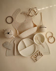 A flat lay of neutral baby feeding items, featuring a confetti-detailed bib, divided plate, bowls, cups, spoons, teethers, rattles, and the mushie Silicone Placemat made from food-grade silicone—all arranged on a light surface.