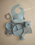 A flat lay of pastel blue and gray mushie baby feeding items—food-grade silicone bib, bowl, divided plate, cup, cat-shaped teether—arranged neatly on a light surface with a mushie Silicone Placemat.