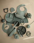 A flat lay of mushie food-grade silicone baby items—bibs, divided plates, spoons, cups with lids and straws, teethers, and Silicone Placemats—displayed in muted green and gray tones on a beige surface.