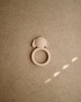 The mushie Dog Teether is a beige silicone teether shaped like a dog’s face with floppy ears, displayed on a light brown textured surface. This non-toxic teether soothes little gums safely and gently.