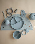A flat lay of light blue food-grade silicone baby feeding items—plate, bowl, handled cup, spoon, fork, teether, rocket toy—plus a Mushie Silicone Placemat with small colorful shapes.