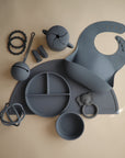 Flat lay of gray mushie silicone baby feeding items, featuring a Snack Cup, bib, divided plate, bowl, mat, teething rings, and pacifier clips—all neatly arranged on a beige surface.