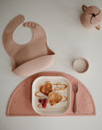 A mushie Silicone Placemat, pink food-grade silicone bib, sippy cup, fork, and cup surround a plate of heart-shaped pancakes with banana slices, raspberries, and syrup on a white surface.