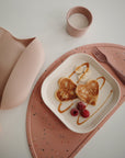 A plate with two heart-shaped pancakes, banana slices, raspberries, and caramel drizzle sits on a pink speckled placemat. Nearby are a pink fork, a mushie Dinnerware Cup with milk, and a pink silicone bib.