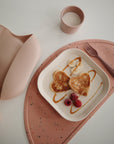 A plate with two heart-shaped pancakes, banana slices, raspberries, and caramel drizzle sits on a pink speckled placemat. Nearby are a pink fork, a mushie Dinnerware Cup with milk, and a pink silicone bib.
