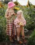 Two young children stand in a sunflower field. One wears a pink hat and plaid dress, holding sunflowers, apples, and a mushie Snack Cup—ideal for toddlers. The other smiles brightly with glasses among the golden blooms.