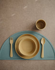 The mushie Dinnerware Cups are shown with a fork, spoon, bowl, and plate in mustard yellow on a teal half-circle placemat, all neatly arranged on a brown surface.