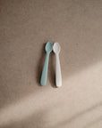 Two mushie Silicone Feeding Spoons, one light blue and one beige, rest side by side on a brown textured surface, with sunlight casting a gentle shadow over them.