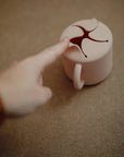 A hand reaches for the flexible, star-shaped opening of the mushie Snack Cup, a spill-proof silicone cup with a handle for toddlers, shown on a brown surface.