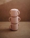 Three small, pink mushie Snack Cups with double handles are stacked vertically on a neutral brown surface and background, creating a minimalist and symmetrical look ideal for toddlers.