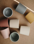 Eight mushie Dinnerware Cups in pink, blue, grey, cream, and mustard yellow are scattered on a brown surface. The non-toxic kids' cups sit at different angles—some upright, others on their sides.