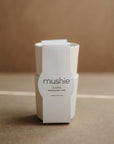 Two stacked cream-colored mushie Dinnerware Cups with a white label reading "mushie CUPS dishwasher safe made in Denmark" rest on a soft brown background. These non-toxic plastic cups are safe for kids and dishwasher friendly.