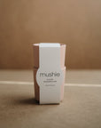 A pair of light pink, non-toxic mushie Dinnerware Cups, dishwasher safe and made in Denmark, are stacked with a white brand band against a brown background.