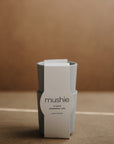 Two stacked light gray "mushie" Dinnerware Cups, each with a white label reading “mushie CUPS dishwasher safe made in Denmark.” Made of non-toxic plastic, these kids cups rest on a neutral light brown surface.