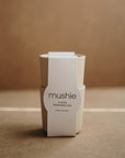 A pair of beige mushie Dinnerware Cups, stacked and wrapped with a white label, are shown against a neutral brown background.