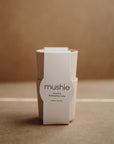 A pair of beige mushie Dinnerware Cups, stacked and wrapped with a white label, are shown against a neutral brown background.