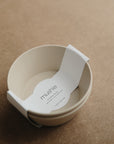 A beige Round Dinnerware Bowl by mushie, made from BPA-free plastic and featuring a white paper label inside, rests on a brown textured surface.