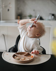 A toddler with curly hair sits in a high chair, tilting their head back while using mushie Silicone Feeding Spoons. A divided plate of food rests on the tray in a bright, modern kitchen.