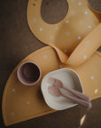 A mushie Silicone Placemat, a sun-patterned silicone baby bib, a small mauve cup, a white bowl, and pink toddler utensils are arranged on a dark surface in warm lighting.