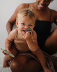 A smiling, light-haired baby in tan holds a teething toy, brush, and FRIGG Daisy Natural Rubber Pacifier. The adult in black sits behind on a soft surface. Shown: FRIGG Daisy Natural Rubber Pacifier 2-Pack by FRIGG.