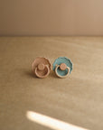 The FRIGG Daisy Silicone Pacifier 2-Pack by FRIGG features two medical grade silicone pacifiers, in muted peach and teal, standing upright on a beige surface with a softly lit beige background.